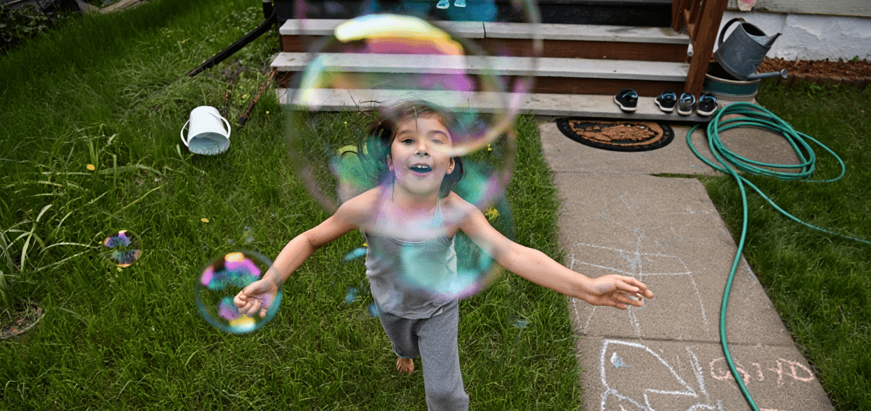 Child running barefoot on grass, smiling, with colorful bubbles floating around them.